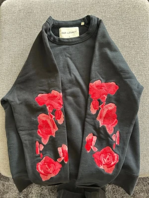 Svart sweatshirt med röda rosor Our Legacy - Säljer en svart sweatshirt från Our Legacy med stora broderade röda rosor på ena ärmen och framsidan. Tröjan har rund halsringning och är tillverkad i mjukt bomullsmaterial. Sälj inte längre 
