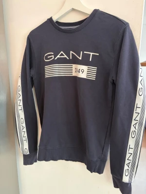Mörkblå sweatshirt från GANT - Snygg mörkblå sweatshirt från GANT med vit logga och tryck på bröstet samt längs båda ärmarna. Tröjan har rund halsringning och ribbade muddar vid ärmslut och nederkant. Perfekt för en avslappnad och trendig look. Passar S
