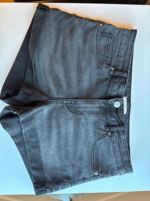 Mörkgrå denimshorts från H&M, storlek 36 - Snygga mörkgrå denimshorts från H&M med klassisk femficksdesign och knappgylf. Shortsen har en midwaist passform och är i perfekt skick. Denimmaterialet har lite stretch.