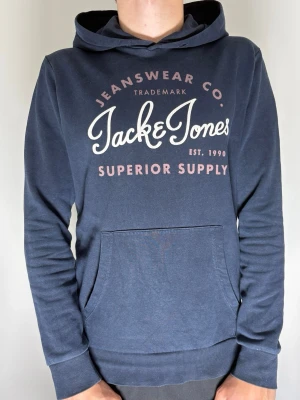 Blå hoodie från Jack & Jones - Tjeeena! Säljer nu denna mörkblåa vintage hoodie från Jack & Jones med tryck framtill i vitt och röd. Klassisk känguruficka och huva. Tillverkad i mjukt bomullsmaterial som är skönt mot huden. Perfekt för en avslappnad och trendig look. 
