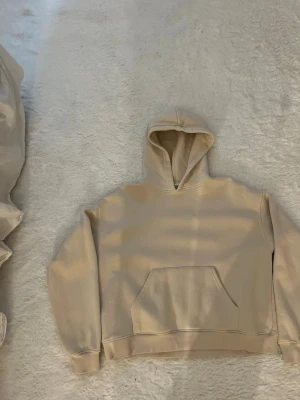 Beige hoodie med huva och magficka - Säljer en stilren beige hoodie med klassisk huva och stor magficka framtill. Hoodien har ribbade muddar vid ärmslut och nederkant. Perfekt för dig som gillar en enkel och clean look. Materialet känns mjukt och skönt mot huden.
