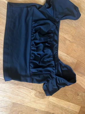 Svart oneshoulder topp från SHEIN - Snygg svart oneshoulder topp från SHEIN med kort ärm och rynkade detaljer framtill. Toppen har en croppad passform och är tillverkad i ett glansigt syntetmaterial som ger en cool look. Perfekt för dig som vill sticka ut lite extra.