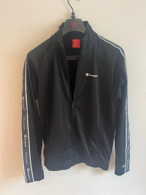 Svart ziptröja från Champion - Svart ziptröja från Champion med vita detaljer och logga på bröstet samt längs ärmarna. Tröjan har hög krage, dragkedja framtill och två fickor. Perfekt för en sportig och avslappnad stil.