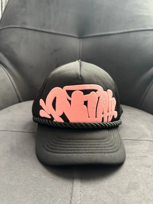 Svart keps med rosa brodyr Synaworld - Säljer en svart truckerkeps från Synaworld med rosa broderad graffiti-text framtill och på skärmen. Kepsen har mesh på sidorna och bak, samt ett svart snöre ovanför skärmen för extra detalj. Justerbar storlek baktill.