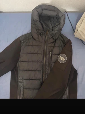 Svart pufferjacka med huva - Säljer en svart pufferjacka med huva och dragkedja. Jackan har quiltad front, mjuka ärmar och en patch med texten 'Supply' på ena ärmen. Praktiska fickor med dragkedja och ribbade muddar. Perfekt för kalla dagar.