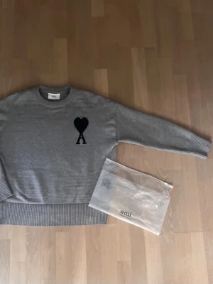 Grå stickad tröja från Ami - Om det är något är det bara att skriva privat . Snygg grå stickad tröja från Ami med svart hjärta och bokstaven A broderat på bröstet. Tröjan har rund halsringning och ribbade muddar vid ärmslut och nederkant. Perfekt för dig som gillar stilrena och trendiga plagg.
