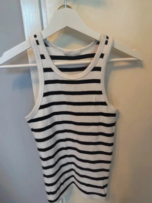 Randig vit och svart linnetopp - Säljer en fräsch linnetopp med svarta och vita ränder. Toppen är ärmlös och har en klassisk rak passform med rund halsringning. Perfekt till jeans eller shorts för en enkel och stilren look. Bra skick!