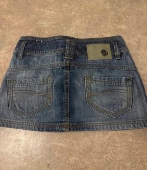 Mini skirt - Säljer en klassisk blå jeanskjol i kort modell med stora bakfickor och bälteshällor. Kjolen har knapp och dragkedja framtill samt detaljerade sömmar. Perfekt för dig som gillar en avslappnad och trendig stil.