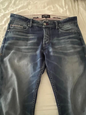 Blå slitna jeans från Replay - Säljer ett par blå jeans från Replay med snyggt slitna detaljer och tvättad look. Klassisk femficksmodell med normal midja och raka ben. Jeansen har vita kontrastsömmar och Replay-logga på insidan av linningen. Perfekta för en avslappnad stil.