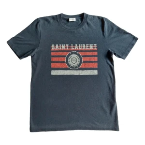 Saint Laurent t-shirt - Snygg mörkgrå t-shirt med tryck från Saint Laurent. Framsidan har vita och röda ränder samt ett coolt emblem i mitten. Klassisk passform och rund halsringning, perfekt till jeans eller shorts.