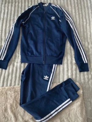 Marinblått Adidas loungewear-set - Snyggt marinblått loungewear-set från Adidas med klassiska vita ränder längs ärmar och ben. Setet består av en zip-jacka och matchande byxor, båda med Adidas logga framtill. Perfekt för chill dagar hemma eller när du vill vara bekväm men ändå se sportig ut.