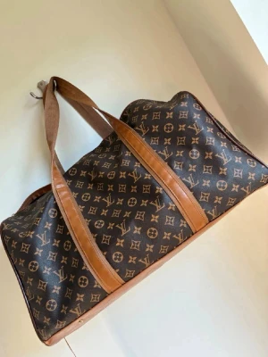 Brun resväska weekendbag  - Snygg brun resväska  med klassiskt monogrammönster och detaljer i ljusbrunt läder. Väskan har två långa handtag och rymlig insida, perfekt för weekendresor. 