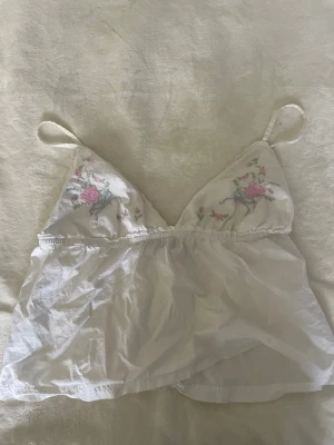 Vit blommig linnetopp med broderi - Söt vit linnetopp med tunna axelband och broderade blommor i rosa och grönt på bröstet. Jättefin att ha till en kofta eller bara som den är.             !pris går att diskutera! 
