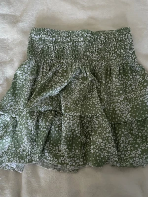 Grön volangkjol med blommigt mönster - Söt kortkjol i ljusgrönt med vitt blommigt mönster och volanger. Kjolen har bred resår i midjan och är gjord i ett lätt, luftigt tyg som passar perfekt till varma dagar. Perfekt för dig som gillar en avslappnad men trendig stil.