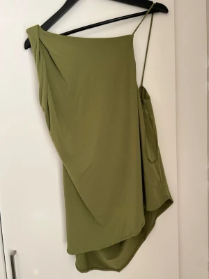 Grönt oneshoulder linne - Säljer ett snyggt grönt linne med asymmetrisk design och oneshoulder-band. Linnet har draperade detaljer och tunna axelband, vilket ger en cool och trendig look. 