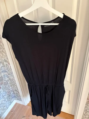 Svart enkel jumpsuit med kort ärm - Snygg svart jumpsuit med korta ärmar och rund halsringning. Midjan har en dragsko för skön passform och ryggen har en liten knappdetalj. Tillverkad i mjukt, tunt tyg som känns bekvämt och luftigt. Perfekt för en avslappnad och trendig look.