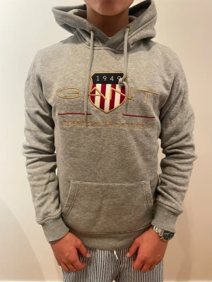 Grå hoodie från Gant - Snygg grå hoodie från Gant med broderad logga och texten 'American Sportswear' på bröstet. Klassisk huva med dragsko och stor magficka framtill. Mjuk bomullsblandning och ribbade muddar vid ärmslut och midja. Perfekt för en avslappnad stil. Storlek S