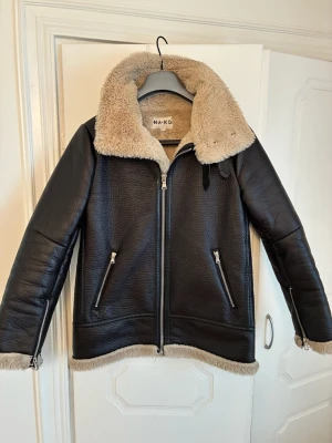Svart skinnjacka från NA-KD med fuskpäls - Snygg svart skinnjacka från NA-KD med beige fuskpäls på krage och insida. Jackan har dragkedja framtill, två fickor med dragkedja och detaljer vid ärmslut. Perfekt för dig som vill ha en trendig och varm look.