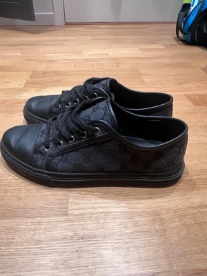Gucci skor - Säljer ett par svarta sneakers med lågt skaft och snyggt monogrammönster på sidan. Skorna har svarta skosnören, metallöljetter och tåparti i läder. Perfekta för dig som gillar stilrena och klassiska sneakers med en twist.