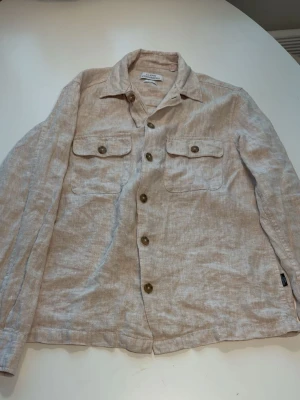 Beige linnéovershirt med fickor - Stilren overshirt i beige linnematerial med två bröstfickor och knappar framtill. Långärmad modell med klassisk krage och knappslå vid ärmslut. Perfekt för lager-på-lager och en avslappnad look.