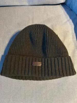 Barbour mössa - Säljer en mörkgrön ribbstickad mössa från Peak Performance. Mössan har uppvikt kant och en liten metallbricka med logga framtill. Perfekt för kalla dagar och enkel att matcha med olika outfits.