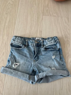 Ljusblå denimshorts med snygga slitningar - Säljer ett par ljusblå denimshorts med upprullade benslut och snygga slitningar framtill. Klassisk femficksmodell med knapp och dragkedja. Perfekta till sommaren för en avslappnad och trendig look.