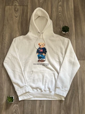 Ralph lauren hoodie med björntryck - Säljer en vit ralph lauren hoodie med huva och känguruficka. På bröstet finns ett tryck av en björn i jeans och blå tröja med amerikansk flagga. Hoodien har ribbade muddar och dragsko i huvan. Perfekt för dig som gillar streetwear och snygga prints.