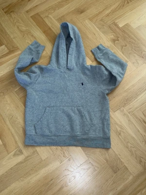 Grå hoodie med huva och magficka. Storlek 10/12 åringar - Snygg grå Ralph Lauren hoodie med klassisk huva och stor magficka framtill. Tröjan har en liten broderad logga på bröstet och ribbade muddar vid ärmar och nederkant. Perfekt för en avslappnad och stilren look.
