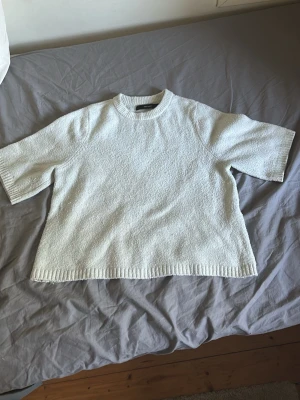 Vit stickad t-shirt med kort ärm - Säljer en vit stickad t-shirt med korta ärmar och rund hals. Tröjan har en avslappnad passform och ribbade kanter vid ärmslut och nederkant. Perfekt för dig som gillar en clean och enkel stil.