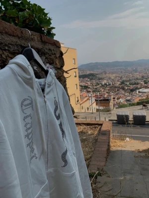 Vit hoodie med ormtryck och stenar - Säljer en vit hoodie med dragkedja och huva. På framsidan finns ett stort ormtryck i svart samt texten trappers i glittrande stenar. Hoodien har fickor framtill och snören i huvan. Perfekt för dig som gillar streetwear och unika detaljer.