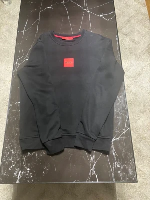 Svart sweatshirt från Hugo - Svart sweatshirt från Hugo med rund halsringning och röd logotyp-patch på bröstet. Tröjan har en klassisk passform med ribbade muddar vid ärmslut och nederkant. Perfekt för en stilren och avslappnad look.