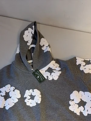 Grå hoodie med vita blommor - Snygg grå hoodie med vita blommönster över hela tröjan. Hoodien har en klassisk huva och är tillverkad i mjukt material som känns skönt mot huden. Perfekt för dig som vill sticka ut med en unik och trendig look.