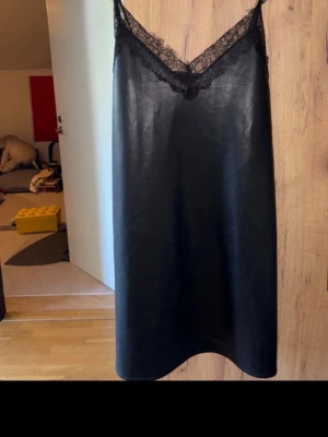 Svart klänning med spetsdetaljer - Stilren svart klänning i syntetmaterial med tunna axelband och v-ringad spetskant upptill. Klänningen har en rak passform och är ärmlös, vilket ger en elegant och enkel look.