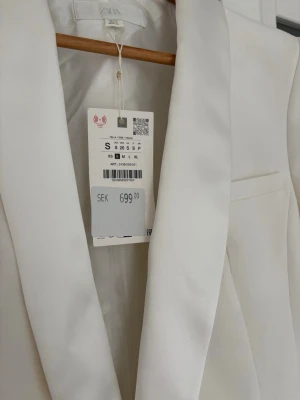Vit smoking kavaj från Zara, storlek S - Stilren vit kavaj från Zara med klassisk krage och en klädd knapp framtill. Kavajen har en slät yta och en bröstficka för en clean look. Perfekt för dig som vill ha en fräsch och modern stil. Kavajen har fortfarande pristagg på . 