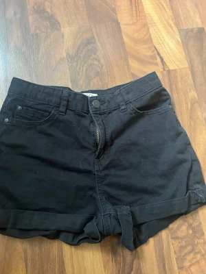 Svarta höga jeansshorts - Svarta shorts i jeansmaterial med hög midja och uppvikta benslut. Klassisk femficksmodell med dragkedja och knapp framtill. Perfekta för varma dagar och enkel att matcha med olika toppar.