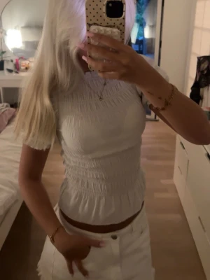 Vit smockad topp med kort ärm - Supersöt vit topp med smockad effekt och korta ärmar. Toppen har en figurnära passform och lätt volang nertill. Perfekt att styla med kjol eller jeans.