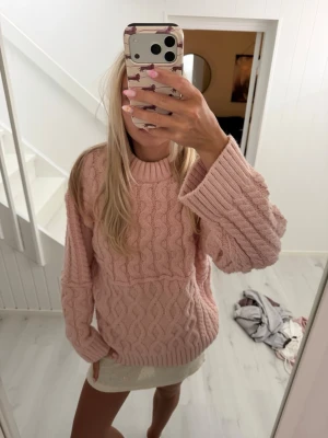 Ljusrosa kabelstickad tröja - Mysig ljusrosa kabelstickad tröja med oversized passform och breda ärmar. Tröjan har ribbade muddar vid ärmslut och nederkant samt en hög, ribbad rund halsringning. Perfekt för att hålla sig varm och snygg under kyliga dagar.