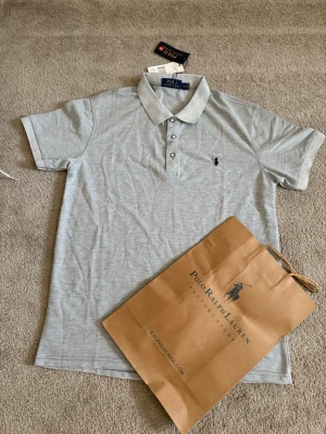 Grå pikétröja från Polo Ralph Lauren - Snygg ljusgrå pikétröja från Polo Ralph Lauren med klassisk krage och knappar framtill. Tröjan har korta ärmar och den ikoniska loggan broderad på bröstet. Perfekt till både jeans och shorts för en clean och stilren look.
