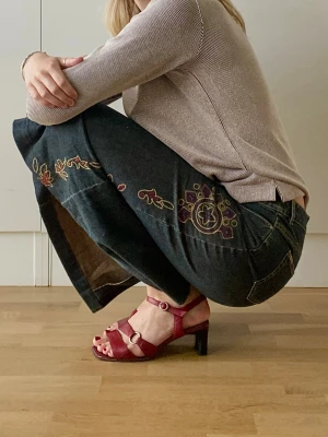 Blommig jeanskjol med broderi - Säljer en mörkblå jeanskjol med broderade blommor i lila, rött och beige längs sidan. Fungerande fickor, knapp och dragkedja fram samt en rak, lång siluett. Vintage och unika detaljer, bra kvalitet och struktur. 