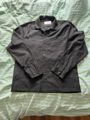Svart overshirt från Woodbird - Sprillans ny svart overshirt från Woodbird med klassisk krage, knäppning framtill och en diskret bröstficka. Den har rutor också. Overshirten har långa ärmar med knapp vid ärmslutningen, mått: midja = 51,5cm längd från axelsömmen t midja = 66cm och ärmen = 66cm skriv om funderingar