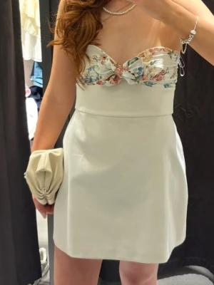 Vit kort klänning med blommig topp - Söt vit kort klänning med axelbandslös design och blommigt mönster upptill. Klänningen har en rosett framtill och en lätt utsvängd kjol. Perfekt för sommarens alla tillfällen.