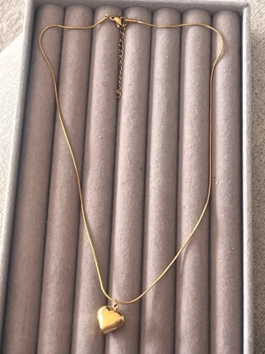 Guldigt halsband med hjärthänge - Snyggt halsband i guldfärgad metall med ett blankt hjärthänge. Kedjan är tunn och elegant med justerbar längd. Perfekt accessoar för att lyfta vilken outfit som helst med en romantisk touch.