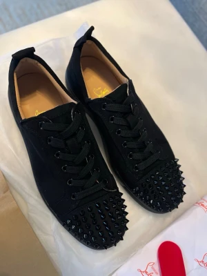 Christian Louboutins med nitar - Säljer ett par svarta Christian Louboutins med coola svarta nitar. Skorna har snörning, låg profil och är tillverkade i mockaliknande material. Perfekta för dig som vill sticka ut med en edgy stil.