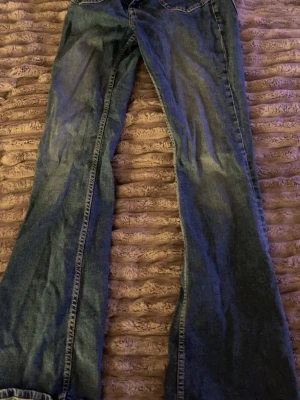 Blå bootcut jeans lågmidjade  - Säljer ett par blå bootcut jeans med snygga slitningar och klassisk femficksdesign. Jeansen har dragkedjegylf och knapp framtill. Materialet är denim i bomull och passformen är något utsvängd nedtill. Perfekta för en avslappnad och trendig look.
