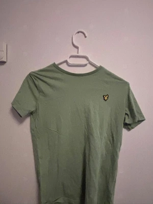 Grön t-shirt från Lyle & Scott - Snygg grön t-shirt från Lyle & Scott med klassisk gul logga på bröstet. T-shirten har rund hals och korta ärmar, perfekt för en avslappnad stil. Tillverkad i mjuk bomull som känns skön mot huden.