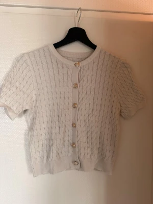 Kortärmad beige kabelstickad kofta - Söt kortärmad kofta i beige med kabelstickat mönster och dekorativa guldfärgade knappar framtill. Koftan har rund halsringning och ribbade kanter. Perfekt att styla över ett linne eller klänning för en trendig look.