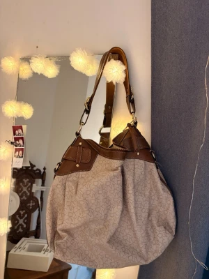 Beige handväska från DKNY - Snygg beige handväska från DKNY med bruna detaljer och guldiga metalldelar. Väskan har ett diskret DKNY-mönster, justerbara axelband och dragkedja upptill. Invändigt finns flera fack för enkel organisering. Perfekt för dig som gillar klassisk och stilren design.