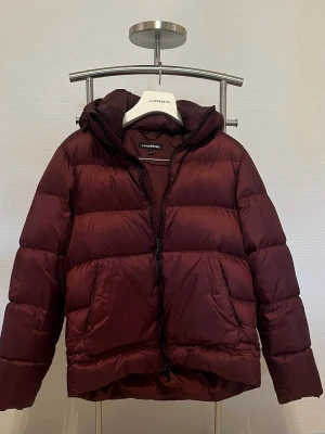 Vinröd pufferjacka från J.Lindeberg - Säljer en vinröd pufferjacka från J.Lindeberg med huva och dragkedja framtill. Perfekt för kalla vinterdagar och riktigt snygg till streetwear-stilen. Den har några små vita prickar framtill som jag inte vet hur de hamnat där men de syns inte tydligt alls. Syns på bild 5! Utöver det är jackan i super bra skick då den bara är använd en halv vinter! Köptes för ca 5000 ordinarie och säljer den nu då jag vill testa något nytt! VID SNABB AFFÄR KAN PRIS DISKUTERAS!