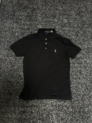 Svart pikétröja från Polo Ralph Lauren - Klassisk svart pikétröja från Polo Ralph Lauren med broderad logga på bröstet. Tröjan har krage, korta ärmar och knäppning med tre knappar framtill. Perfekt för en stilren och avslappnad look.