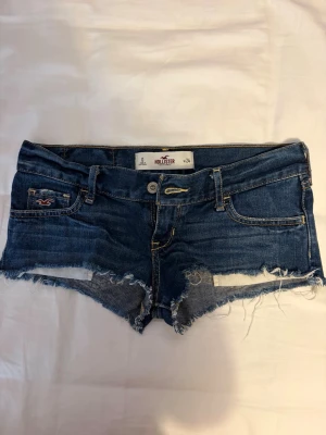 Blå jeansshorts från Hollister - Säljer ett par blå jeansshorts från Hollister med råa kanter och slitna detaljer. Klassisk femficksmodell med knapp och dragkedja framtill. Perfekta för varma dagar och ger en avslappnad look.